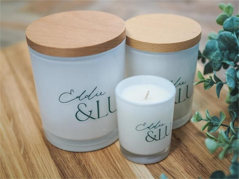 EL cocowax candle trio - white jars, two largest lidded-wooden, on wood-grain table with fresh leaves.
