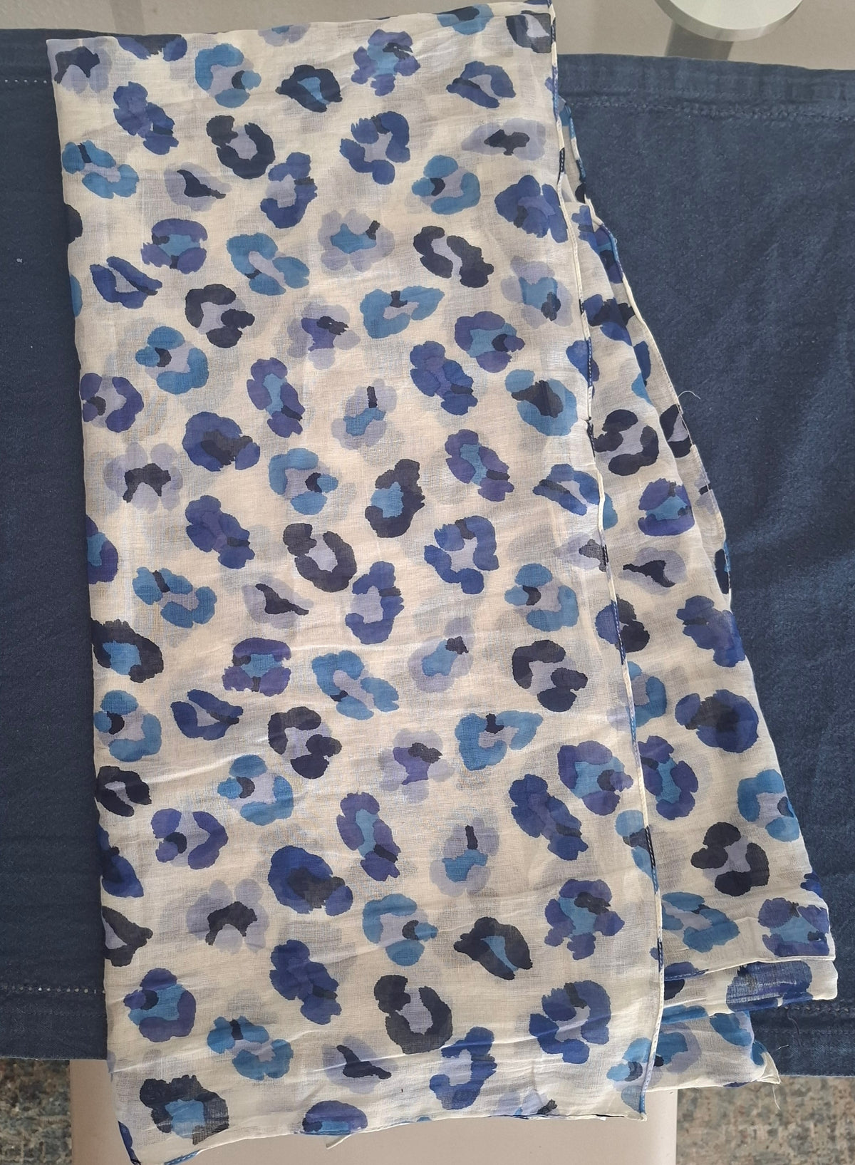 A lightweight white scarf with an all-over blue leopard print pattern, laid flat on top of a dark blue fabric surface.