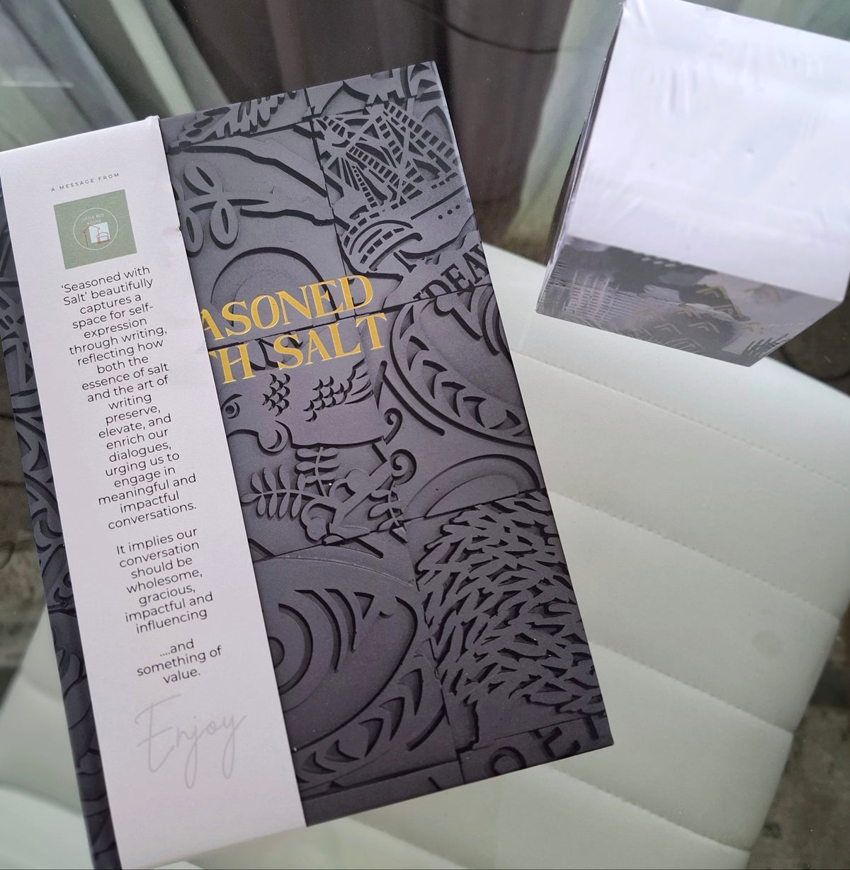 An A5 journal labeled 'Seasoned Salt' with ornate dark gray artwork and a white vertical label, with a memo cube, sides surrounded by artwork, photographed on a white chair.