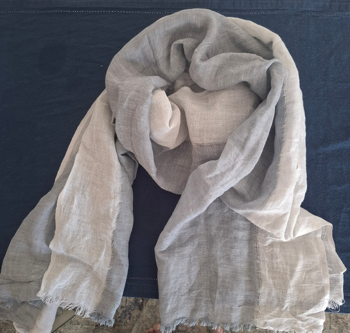 A light beige linen scarf with frayed edges, draped over dark blue fabric.