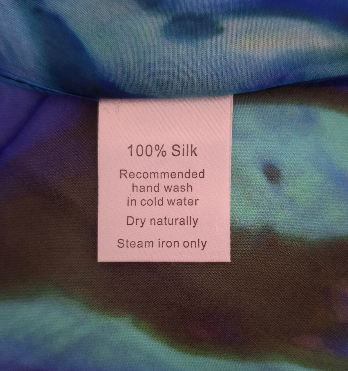 A close-up of a light pink fabric tag reading '100% Silk Recommended hand wash in cold water Dry naturally Steam iron only' attached to a blue and purple silk garment.