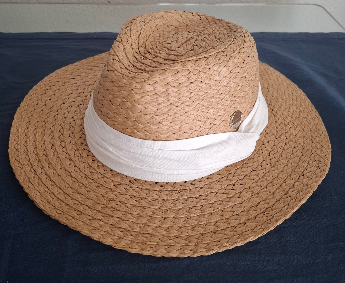 A natural woven straw sun hat with a wide brim and crown, featuring a white fabric band around the middle and a small metallic button detail, displayed on a dark navy blue fabric surface.
