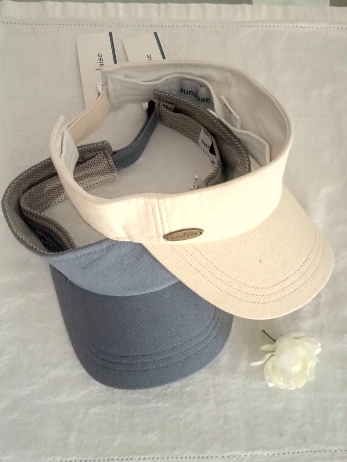 Two visors, one beige and one gray, on a textured surface with a white flower.