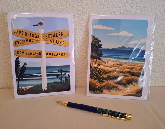 Two retro-vintage-style illustrated cards (in clear sleeves), displayed on a beige surface with a blue and gold and paua  pen in front. The left card shows a coastal signpost with New Zealand place names, and the right card shows a beach and ocean landscape.