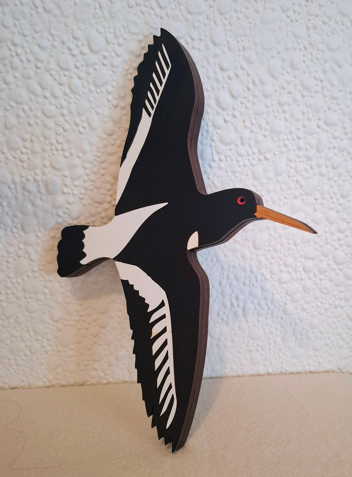A stylized wooden wall hanging of a flying bird with black and white body and wings, a small red eye, and a long orange beak, shown against a textured off-white wall.