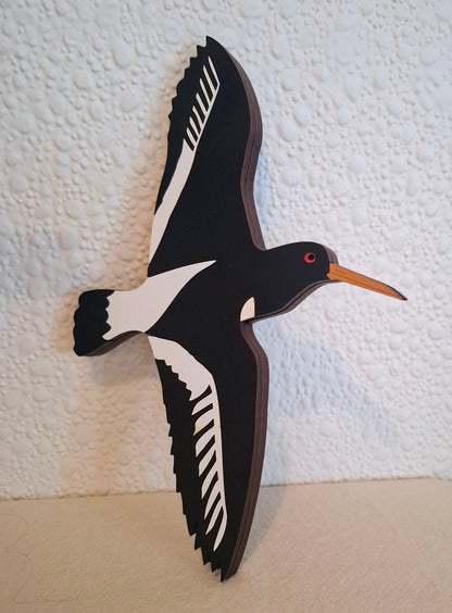 A stylized wooden wall hanging of a flying bird with black and white body and wings, a small red eye, and a long orange beak, shown against a textured off-white wall.