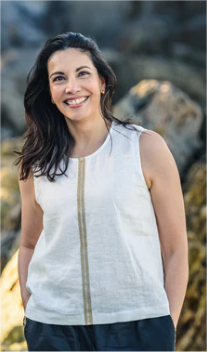 Woman wearing a sleeveless white top with gold trim standing against a natural background.