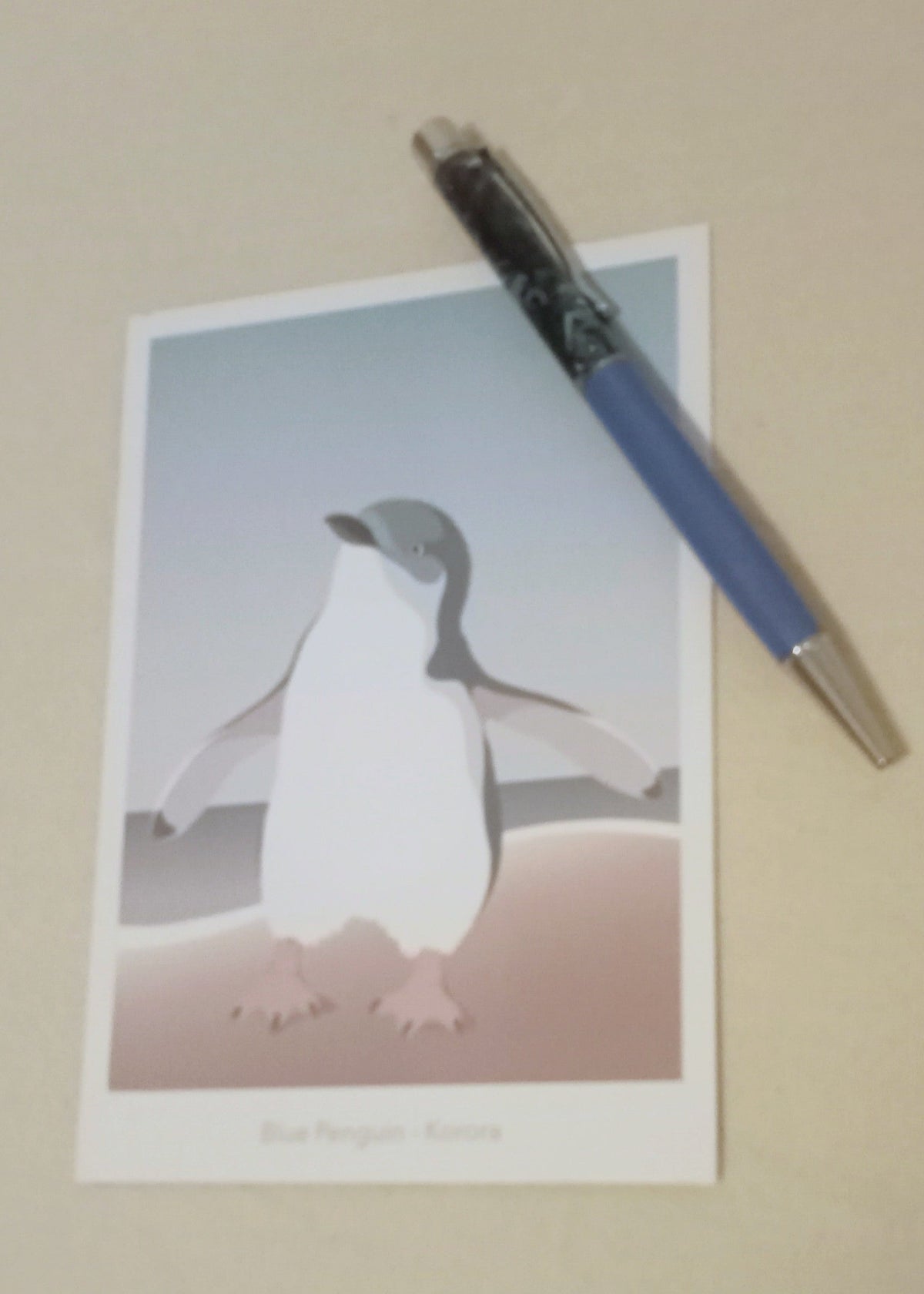 Postcard with a blue penguin illustration and a paua pen on a beige background