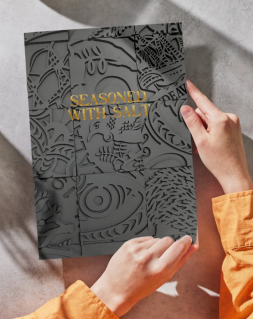 Purple-grey A5 suede-looking journal with pattern etched on cover and words "seasoned with Salt" embossed in gold on front - held in hands emiting from orange shirt, journal set on white background, presumed table.