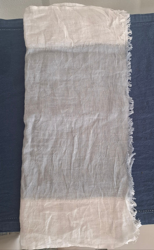 A rectangular white woven fabric scarf  with frayed edges, laid flat on top of dark blue fabric.