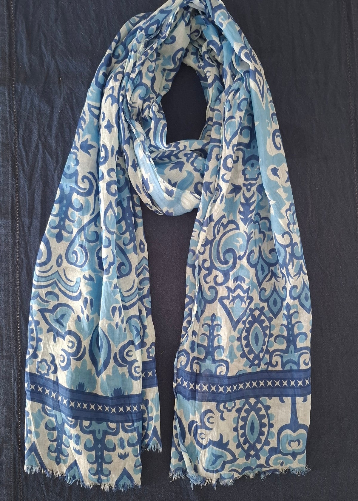 A lightweight scarf with a blue and white traditional pattern, laid flat on a dark navy blue fabric surface.
