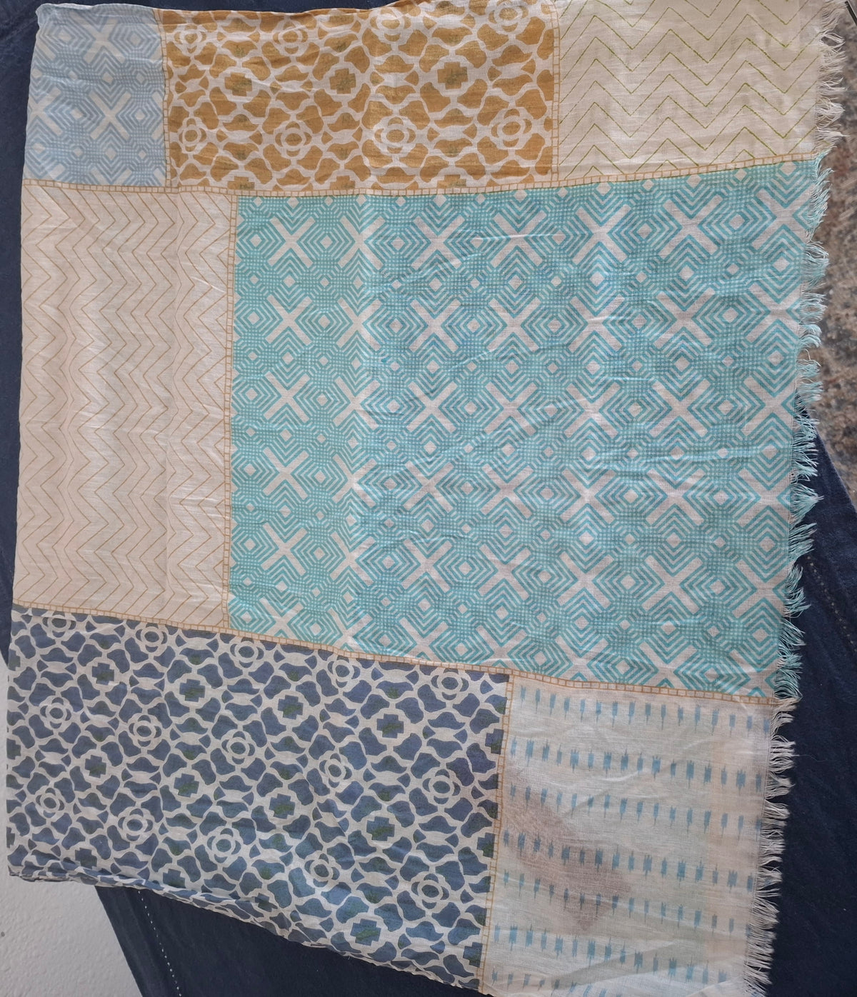 A square patchwork scarf laid flat on a dark surface, featuring multiple panels in light blue, beige, and navy with geometric and chevron patterns and fringed edges.