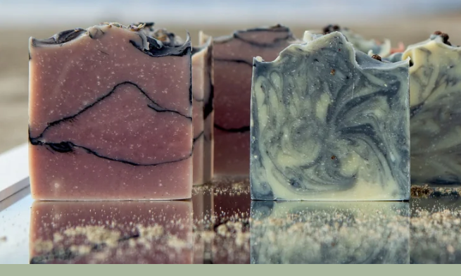 Several artisanal marbled soap bars are arranged upright on a reflective surface. The front soaps show swirled patterns in dusty pink, and blue+cream, with small speckled inclusions throughout.
