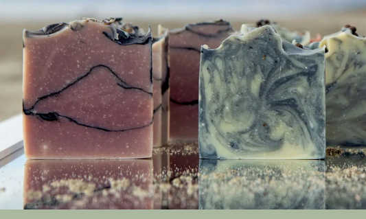 Several artisanal marbled soap bars are arranged upright on a reflective surface. The front soaps show swirled patterns in dusty pink, and blue+cream, with small speckled inclusions throughout.