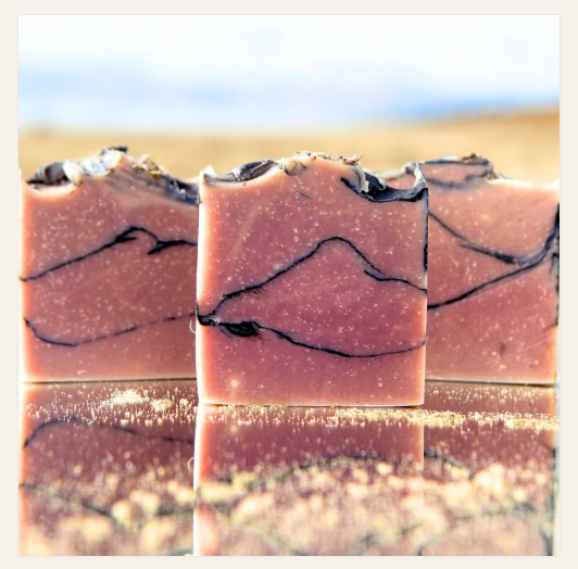 Three rectangular bars of handmade soap with a dusty rose pink color and black marbled swirls, standing on a reflective surface with a blurred beach background.