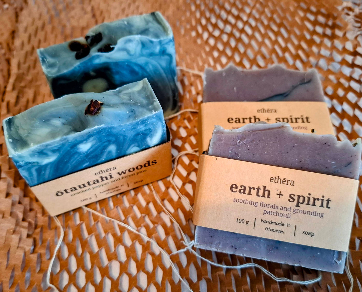Four handmade artisan soaps from the brand Ethēra, displayed on a woven wicker surface. The set includes two blue and white marbled soaps with cracked peppercorns, and two lavender-purple soaps, each bar wrapped in kraft paper sheaths labeled 'Ethēra' plus fragrance type.