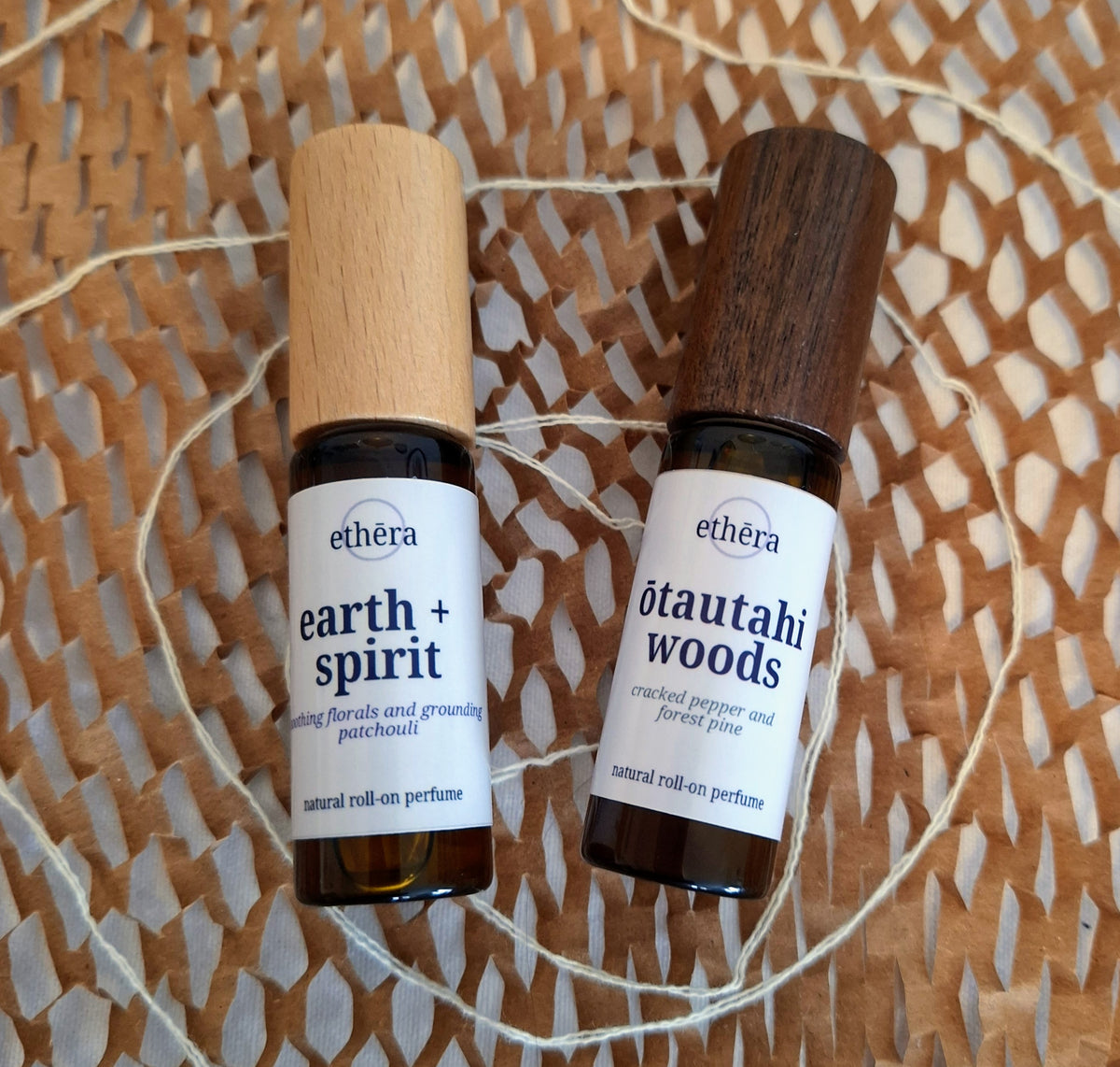 Two amber glass roll-on perfume bottles with white labels from the brand ethéra, placed on a woven brown paper mat with white string. One bottle has a light wood cap and is labeled 'earth + spirit', and the other has a dark wood cap and is labeled 'ōtautahi woods'.