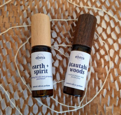 Two amber glass roll-on perfume bottles with white labels from the brand ethéra, placed on a woven brown paper mat with white string. One bottle has a light wood cap and is labeled 'earth + spirit', and the other has a dark wood cap and is labeled 'ōtautahi woods'.