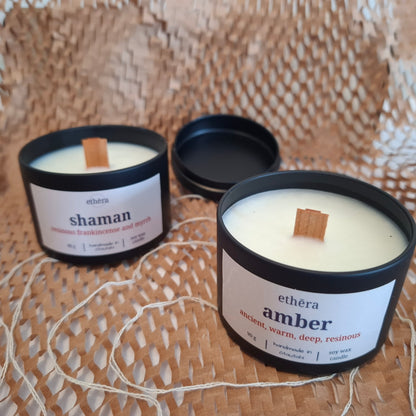 Two white soy candles in matte black jars with white labels, shown on a woven straw surface. One label reads 'ethéra shaman' and the other 'ethéra amber'; both have wooden wicks, and one jar is open with its lid placed beside it.