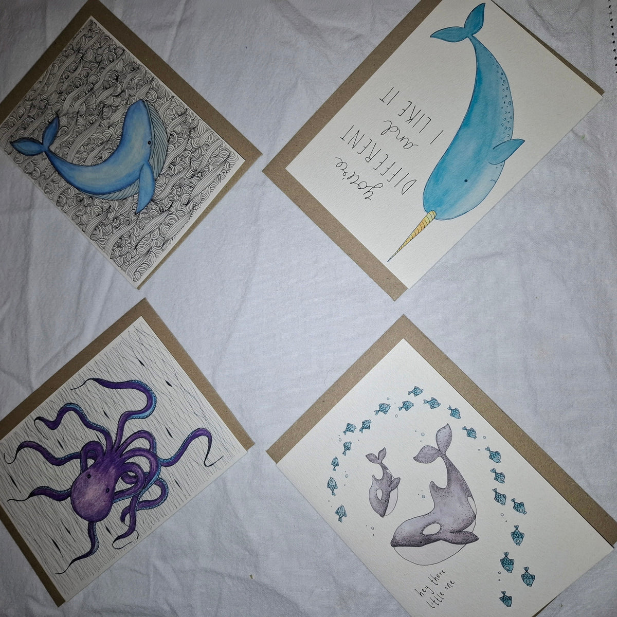 Four handmade art cards featuring marine life illustrations, including a blue whale, narwhal, purple octopus, and gray whale with fish, arranged on a white fabric background.