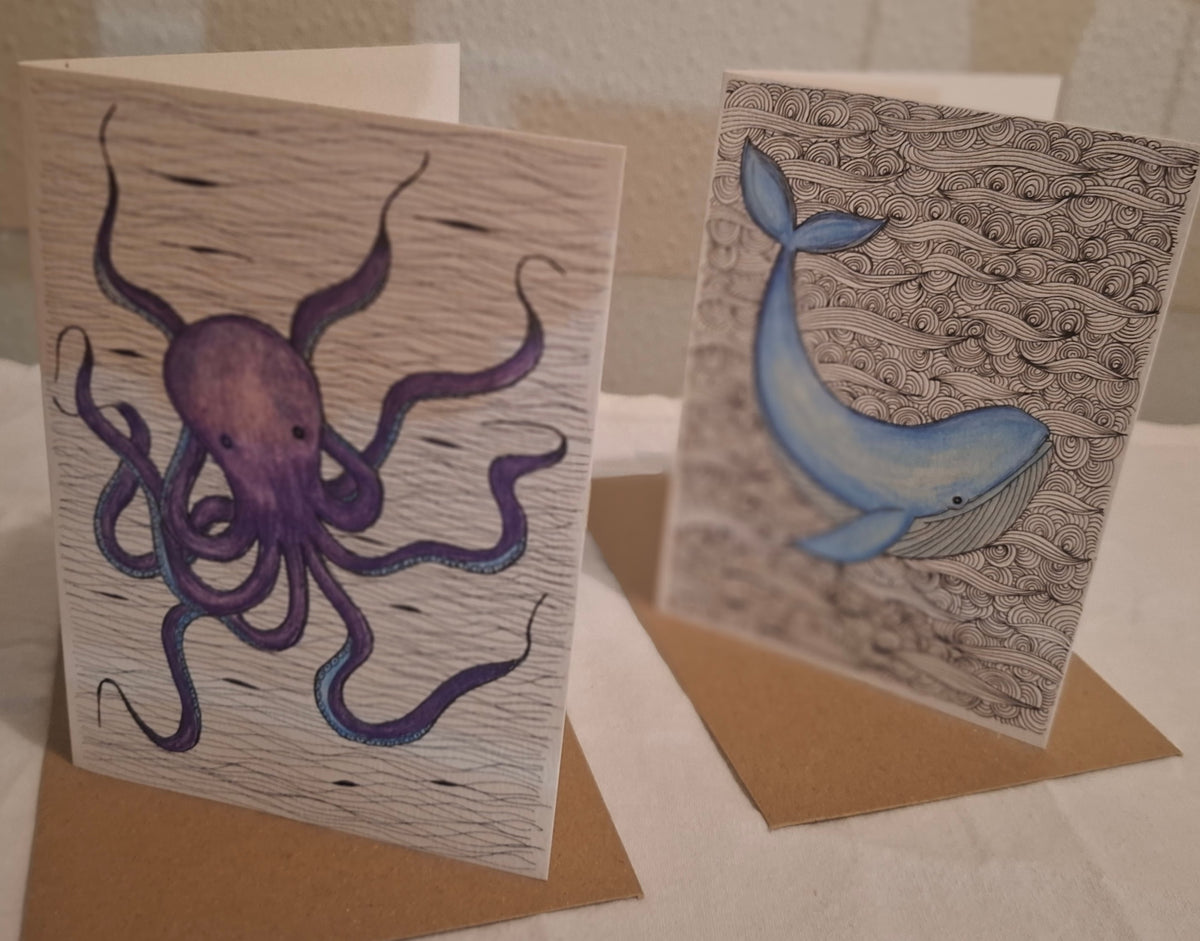 Two handmade greeting cards with ocean-themed illustrations, standing upright on brown kraft envelopes. The left card features a purple octopus on a wood-grain textured background, and the right card features a blue whale on a black-and-white wave patterned background.