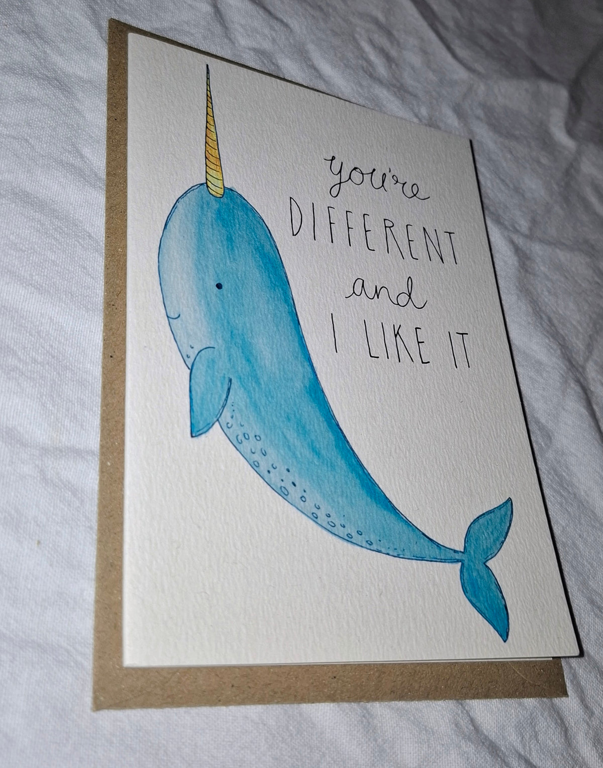 A handmade greeting card featuring a watercolor illustration of a blue narwhal with a gold horn and the handwritten text 'you're DIFFERENT and I LIKE IT'. The card is placed on top of a brown kraft envelope on a wrinkled white fabric background.