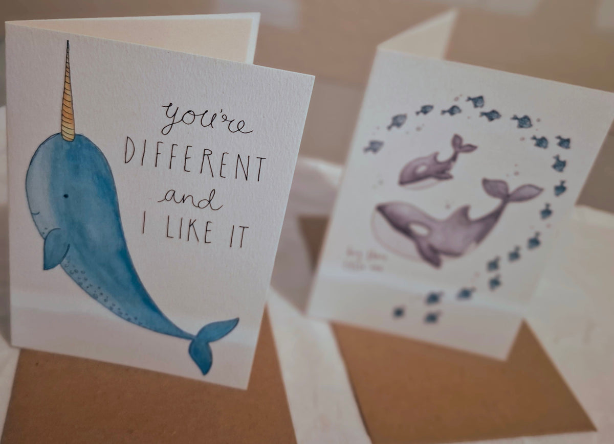 Two handmade greeting cards displayed on brown kraft envelopes. The card in the foreground features a watercolor illustration of a blue narwhal with a gold horn and the text 'you're DIFFERENT and I LIKE IT'.