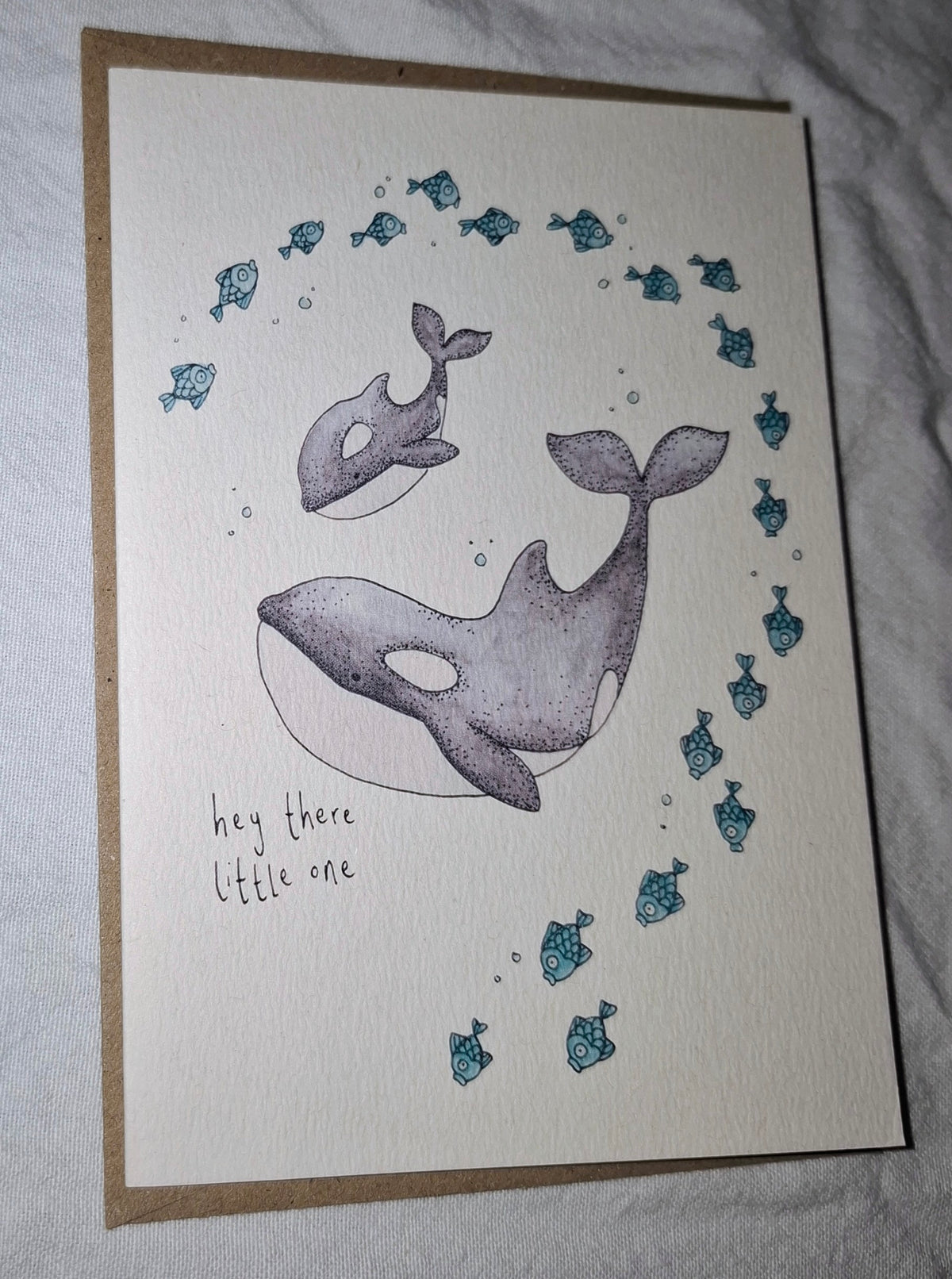 A handmade greeting card featuring a watercolor illustration of an orca and a smaller whale, surrounded by small blue fish and bubbles, with the handwritten text 'hey there little one'. The card is placed on a brown kraft envelope against a wrinkled white fabric background.