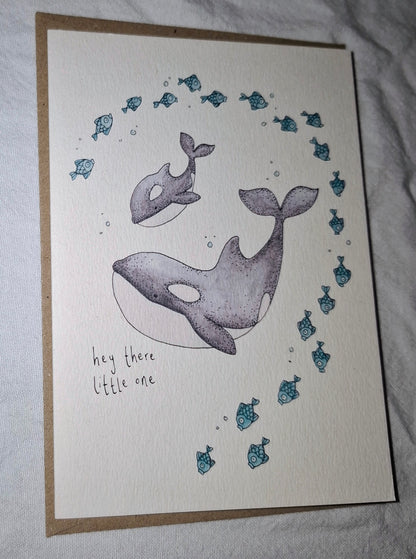 A handmade greeting card featuring a watercolor illustration of an orca and a smaller whale, surrounded by small blue fish and bubbles, with the handwritten text 'hey there little one'. The card is placed on a brown kraft envelope against a wrinkled white fabric background.
