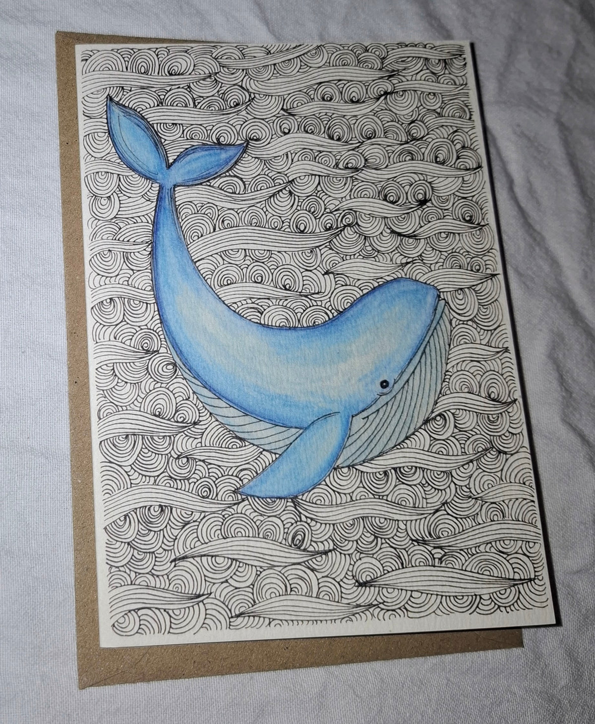 A square art print or card featuring a blue whale illustration swimming over black-and-white wave patterns, placed on top of a brown kraft envelope against a light fabric background.