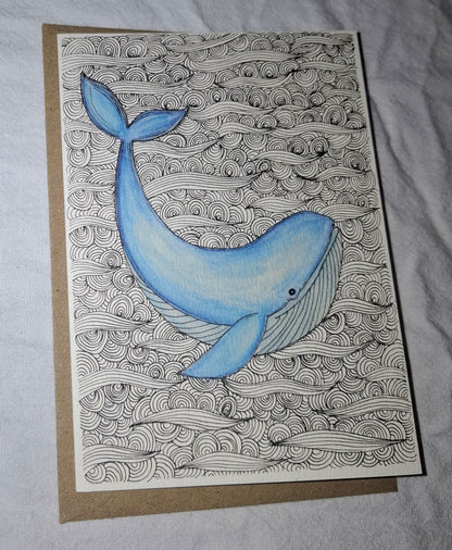 A square art print or card featuring a blue whale illustration swimming over black-and-white wave patterns, placed on top of a brown kraft envelope against a light fabric background.