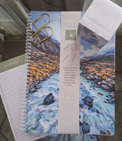 A spiral-bound 2027 planner with a scenic mountain river cover, shown with a to-do list pad, gold paperclips, and a white-paper art-memo-cube on a table.