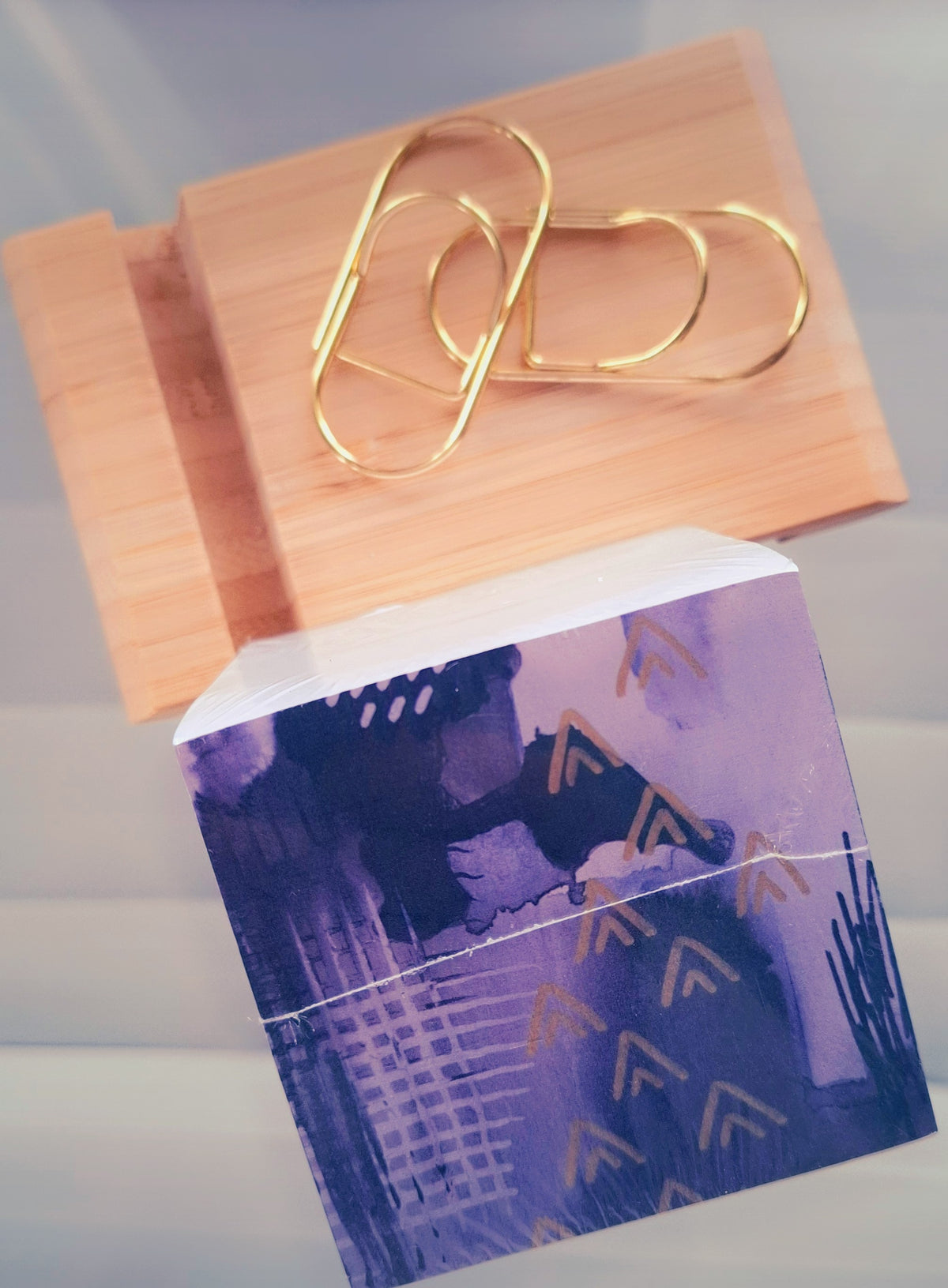 A close-up of a purple and blue patterned memo cube block with gold foil chevron accents, placed on top of a light wood block that holds two gold pen-clips, on glass background.