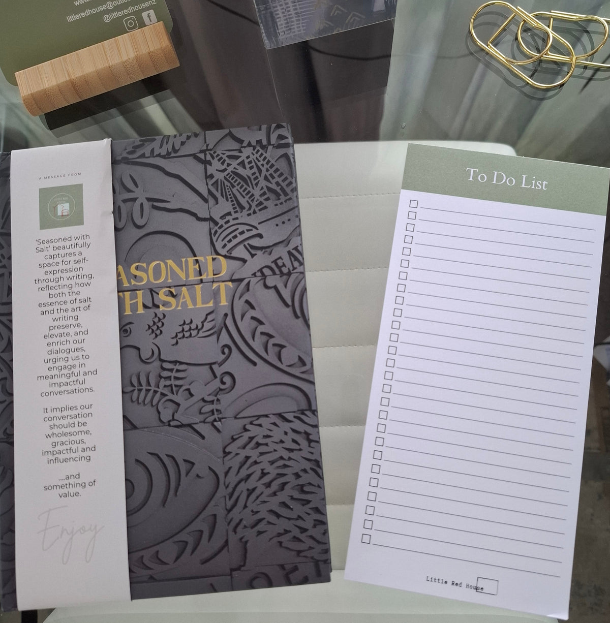 A close-up of a branded stationery set - a dark gray journal with embossed floral detailing and gold text reading 'Seasoned with Salt, a white to-do list pad with a sage green header, a pair of gold pen-clips - and a wooden mobile holder containing a business card with printed text. 
