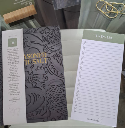 A close-up of a branded stationery set - a dark gray journal with embossed floral detailing and gold text reading 'Seasoned with Salt, a white to-do list pad with a sage green header, a pair of gold pen-clips - and a wooden mobile holder containing a business card with printed text. 