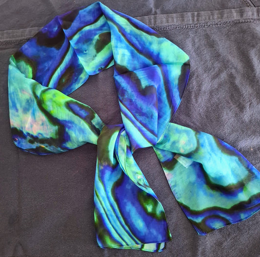 A lightweight scarf with an abstract, marbled pattern in shades of blue, green, and purple, tied in a knot and laid flat on a textured gray fabric background.