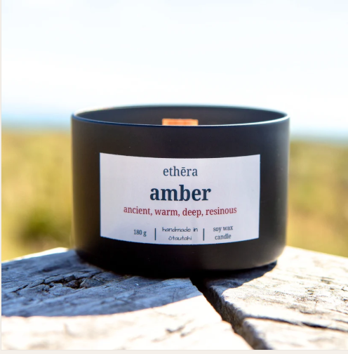 White soy candle in black jar with 'ethera amber' label on a wooden surface with soft focus natural background